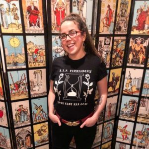 Picture of Kate Sheridan, a.k.a The Laundress. She standing in front of a folding screen of tarot cards. She is wearing black jeans with a red belt. She has a black shirt on that says "R.I.P. Patriarchy" It has two white roses and an hourglass timer on the shirt. She is smiling with her hands in her front pockets.